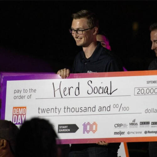 A group of people smile and celebrate as two men hold a large presentation check made out to "Herd Social" for \$20,000. The check is from Demo Day 2024, with logos from sponsors like Meijer, PNC Bank, and Huntington visible at the bottom.
